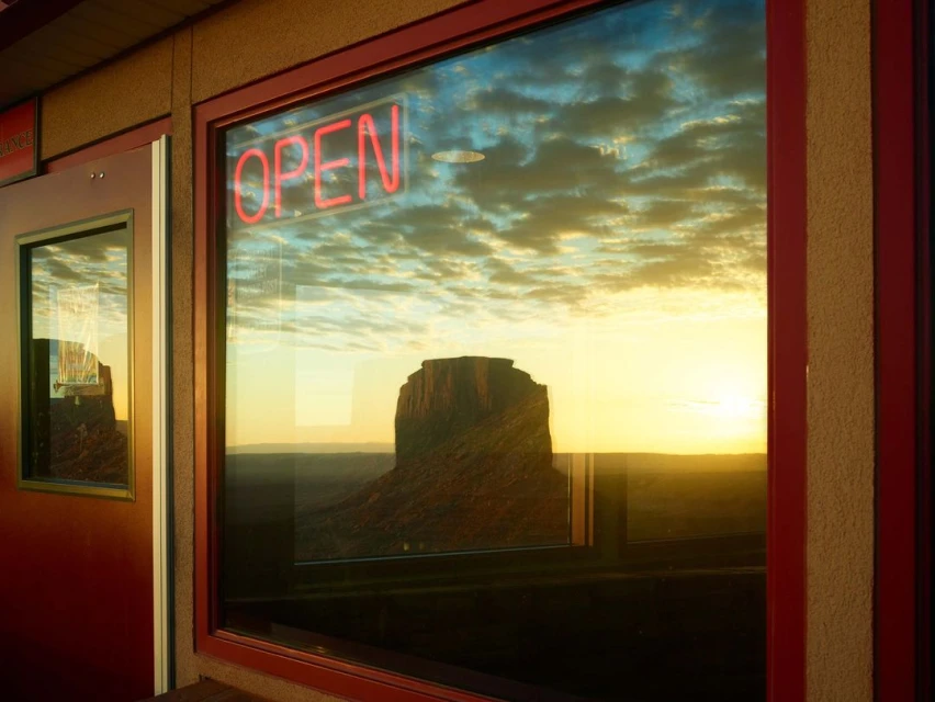 Monument Valley vista reflected in a window with a red neon 'OPEN' sign. Sunset sky and clouds add dramatic atmosphere.