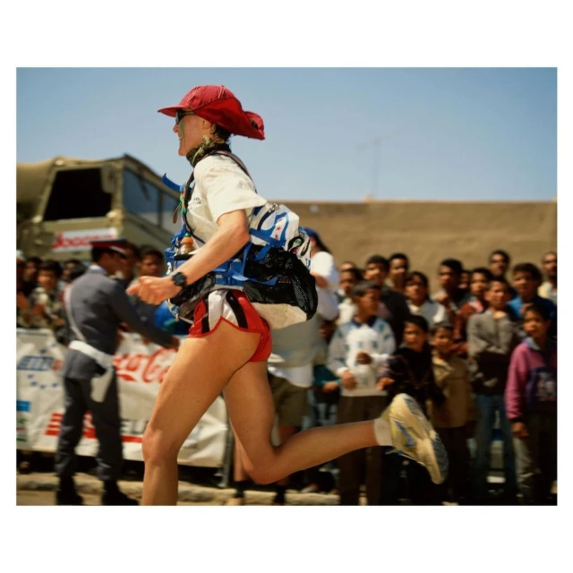 Woman runner competing outdoors, wearing red hat and running shorts, with a crowd watching and a Coca-Cola banner in the background.