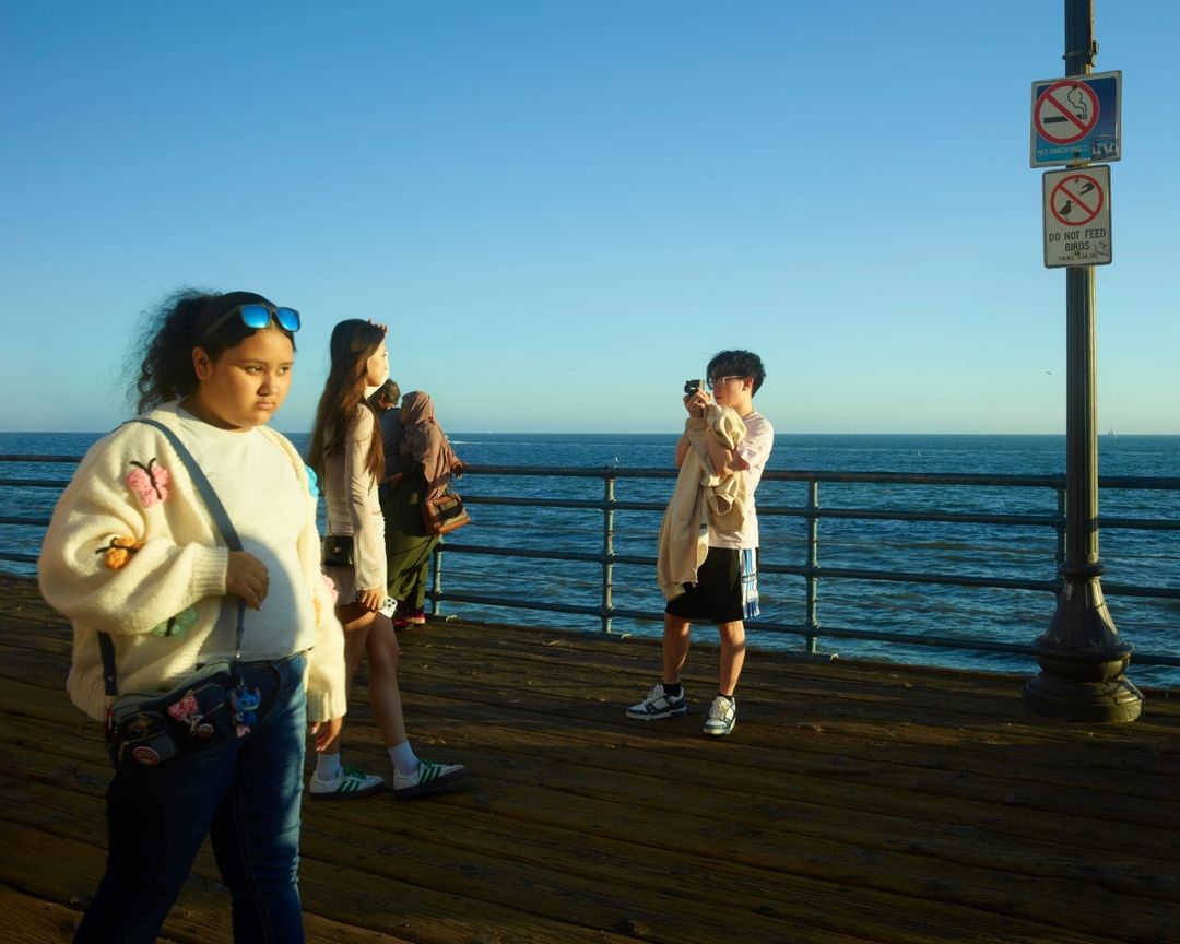 Three people on a pier next to the ocean, including a young girl with butterfly-themed sweater walking in the foreground during the day.