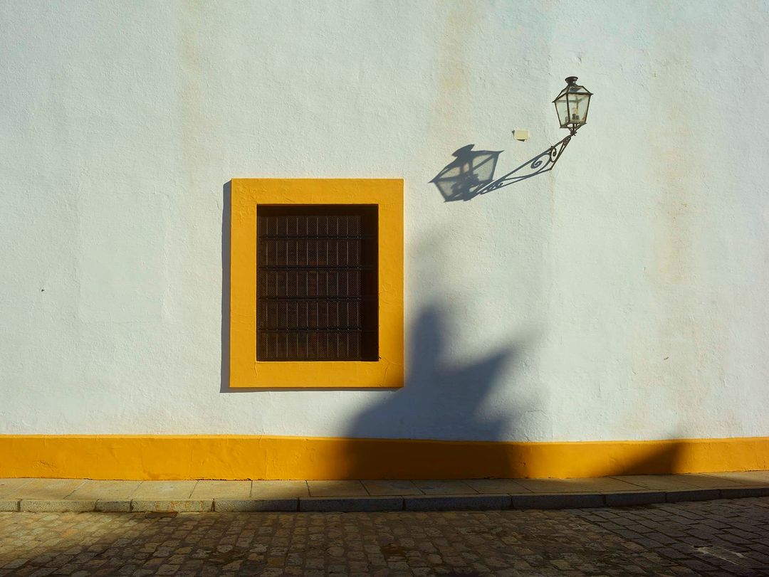 Exterior of a building with a window with yellow trim and a street lamp casting shadows on the wall, creating an abstract visual.