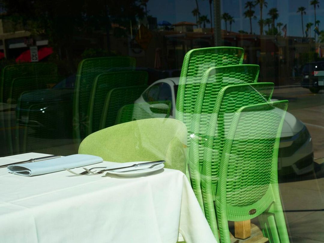 Outdoor dining scene with a set table and stacked green chairs reflecting the blurred palm trees and buildings of the city.