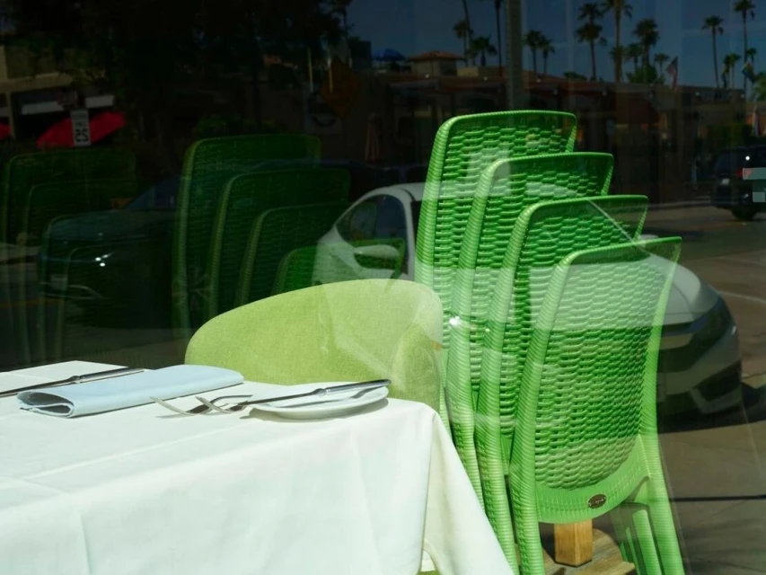 Outdoor dining scene with a set table and stacked green chairs reflecting the blurred palm trees and buildings of the city.