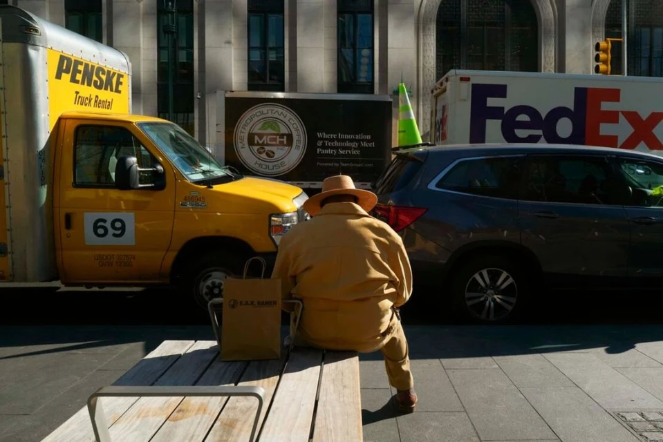 An adult wearing a hat and tan outfit sitting on a bench next to a yellow Penske truck and brown LAX Samir shopping bag in an urban setting.