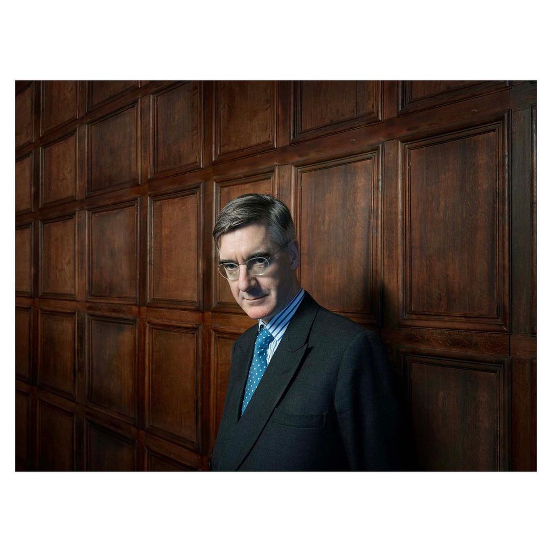 Portrait of an adult male in a dark suit and glasses, standing indoors against a dark paneled wood wall, with a medium shot.