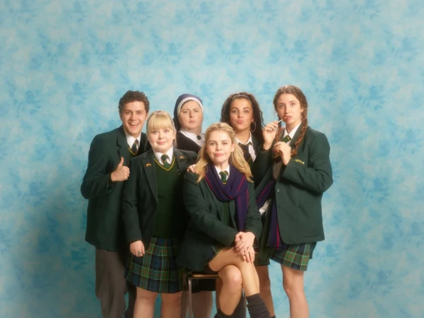 Group of six young adults and one adult woman in a nun's habit pose in school uniforms against a blue, mottled studio backdrop.