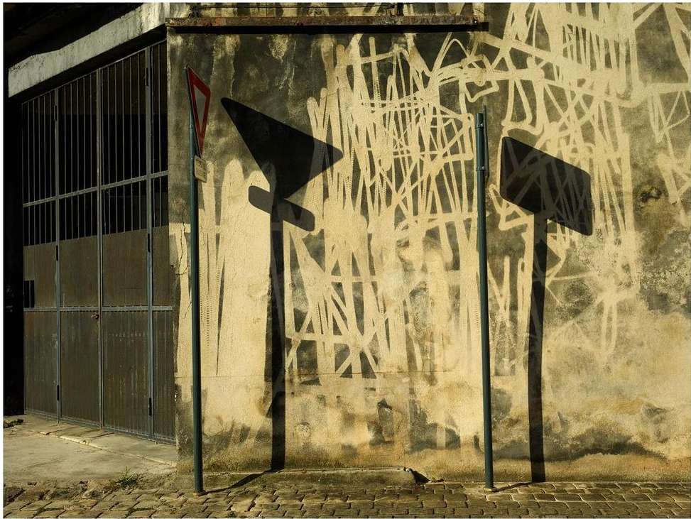Outdoor shot of two street signs throwing a shadow on a weathered building with graffiti near a gated entrance on a brick street.