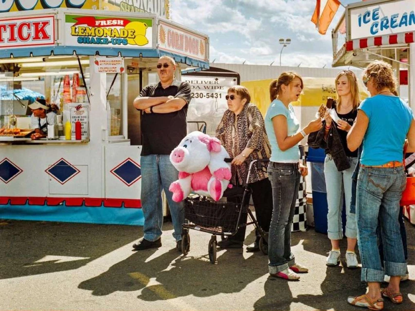 Outdoor fair scene with a group of people standing near food stands, featuring adults, teenagers, and a plush pig in a cart.