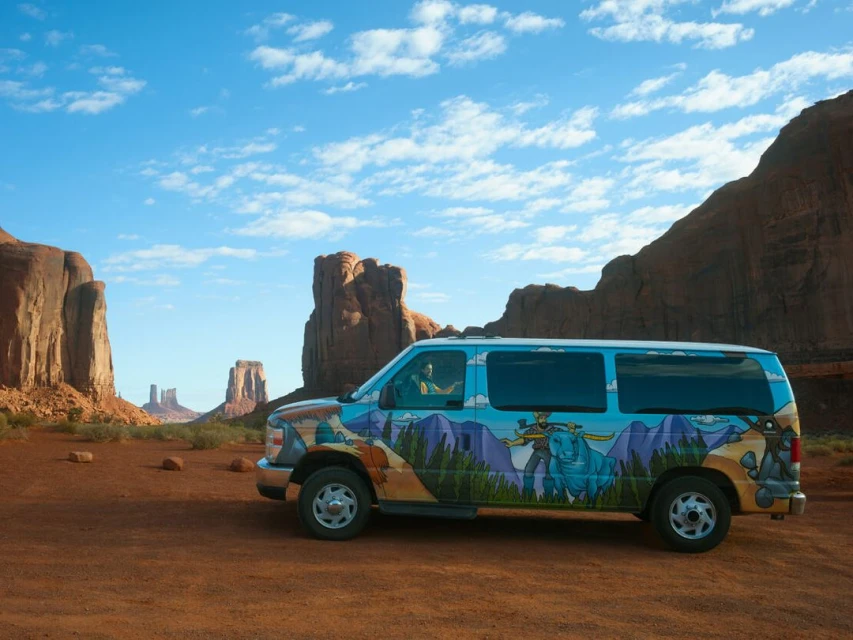 Painted van parked in Monument Valley, Utah, with towering sandstone buttes under a blue sky with scattered clouds, showcasing desert scenery.