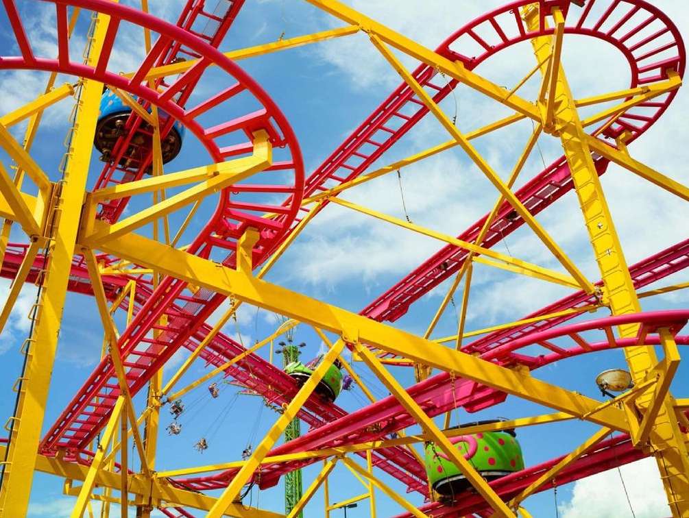 Amusement park ride featuring a yellow framework and red tracks set against a blue sky with scattered clouds; riders visible.