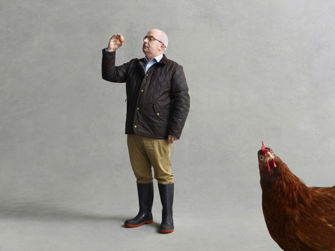 Man wearing a quilted brown jacket and rubber boots inspecting an egg, with a large chicken visible in the foreground corner.
