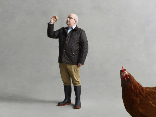Man wearing a quilted brown jacket and rubber boots inspecting an egg, with a large chicken visible in the foreground corner.