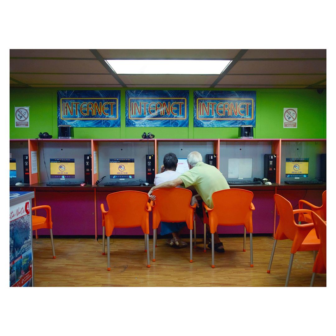 Two elderly people looking at a computer screen in an internet cafe with orange chairs and lime green walls, lit by fluorescent lights.