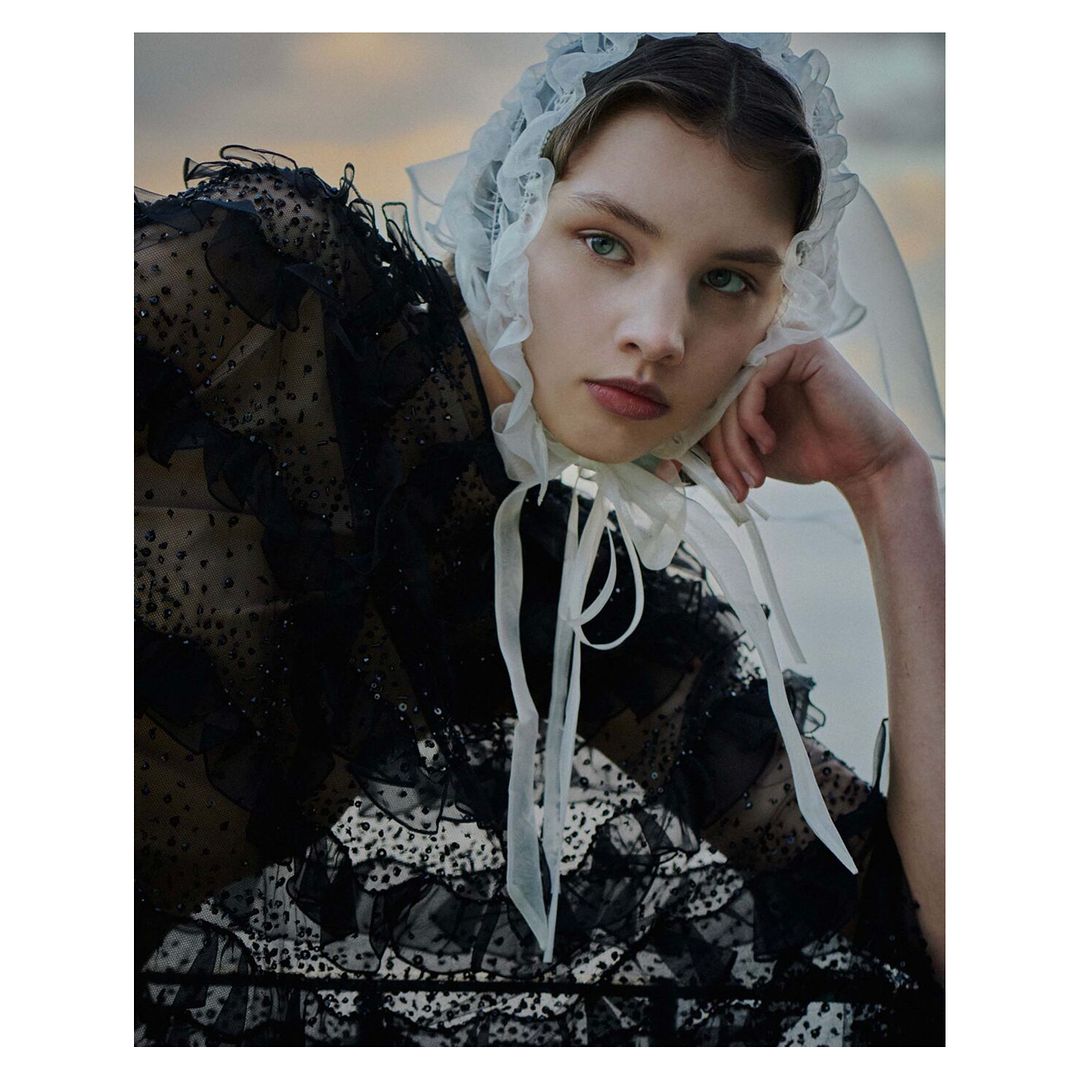 Close-up portrait of a young woman with fair skin and light eyes, wearing a black, polka-dot tulle gown with a white lace bonnet.
