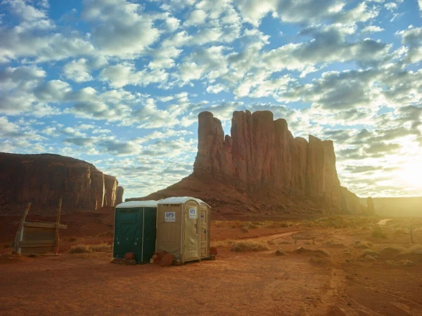 Daytime view of the red rock formations of Monument Valley, Arizona, with a blue sky, scattered clouds, and two portable restrooms.