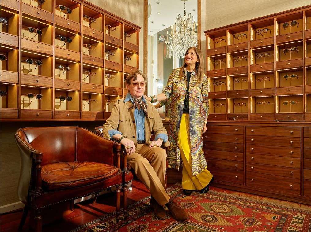 Two adults in a vintage eyeglass store, surrounded by wooden eyewear displays and a plush chair, creating a retro-chic atmosphere.