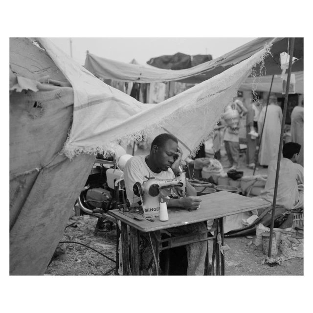 Monochrome shot of a man using a vintage Singer sewing machine at an outdoor makeshift workshop with blurred figures in background.