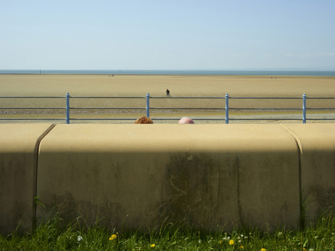 Two people's heads barely visible behind a concrete wall overlooking a wide sandy beach and calm ocean by Dan Burn-forti