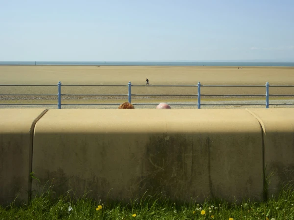 Two people's heads barely visible behind a concrete wall overlooking a wide sandy beach and calm ocean by Dan Burn-forti
