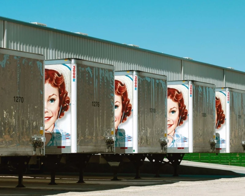 A row of trailers with a vintage graphic of a smiling woman, parked near a grey warehouse against a clear blue sky backdrop.