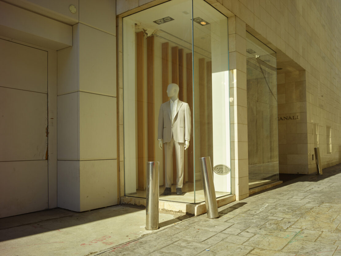 Mannequin in a beige suit stands in a shop window with glass walls on a city street with stone pavement and light-colored buildings.