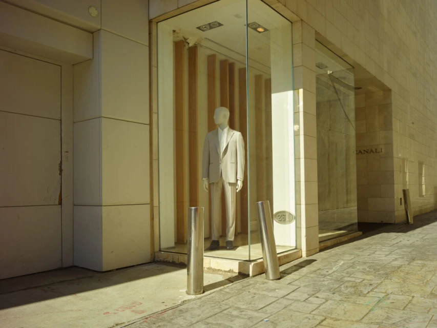 Mannequin in a beige suit stands in a shop window with glass walls on a city street with stone pavement and light-colored buildings.