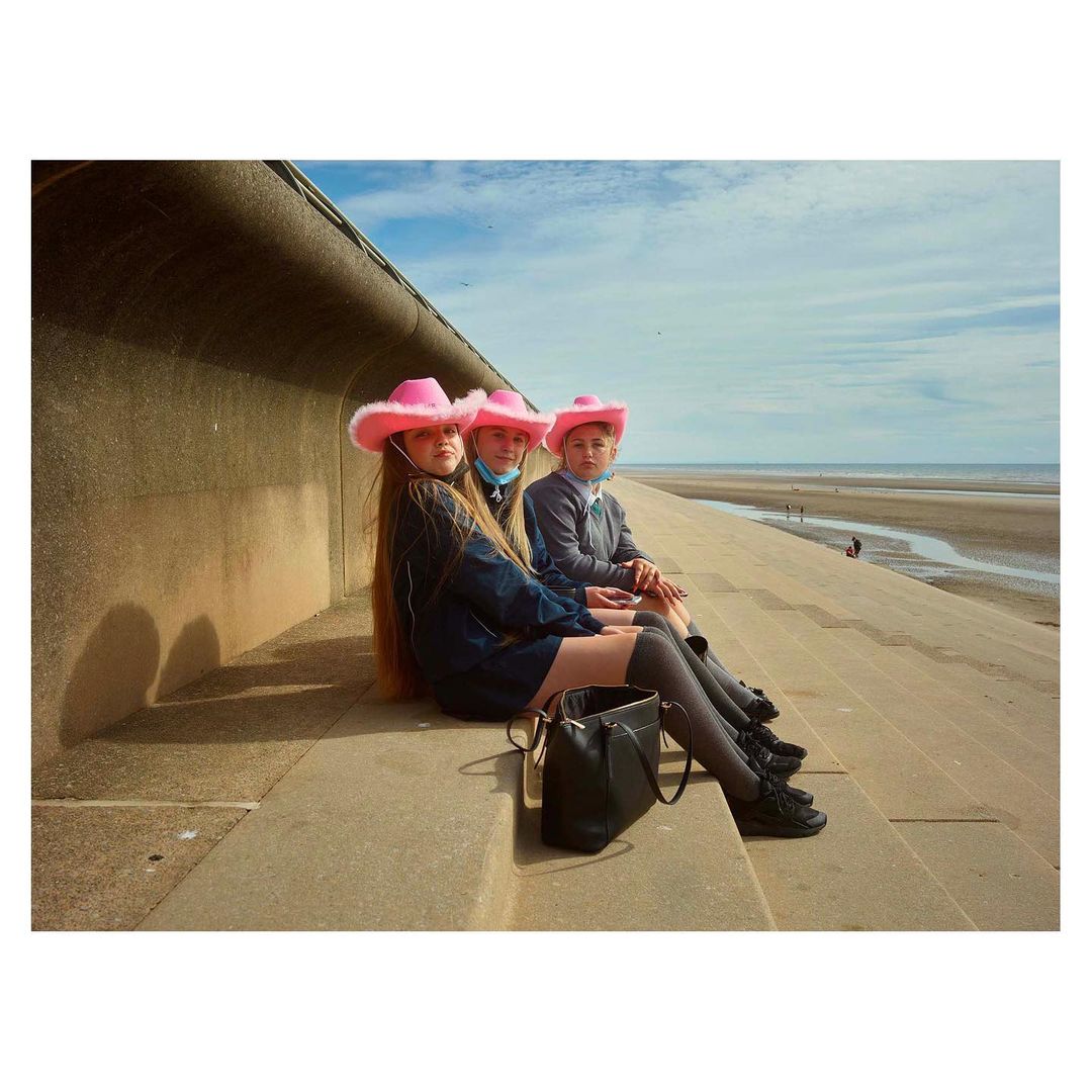 Three young people in pink cowboy hats sit on concrete steps overlooking a beach on a sunny day, showcasing fashion and leisure.