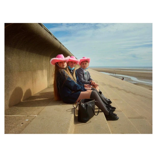 Three young people in pink cowboy hats sit on concrete steps overlooking a beach on a sunny day, showcasing fashion and leisure.
