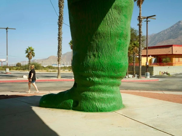 A large green fiberglass boot roadside attraction, with a woman walking on the sidewalk nearby on a sunny day in an outdoor environment.