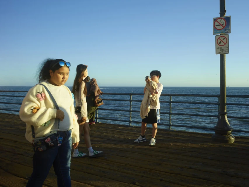 Three people stand on a pier on a sunny day. One woman walks toward the camera wearing jeans and a butterfly sweater.