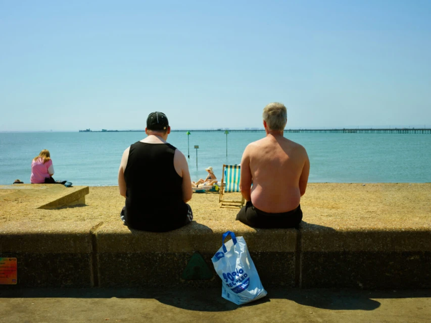 Two people sit on a concrete wall overlooking the ocean on a sunny day with a beach in the background and a blue bag.