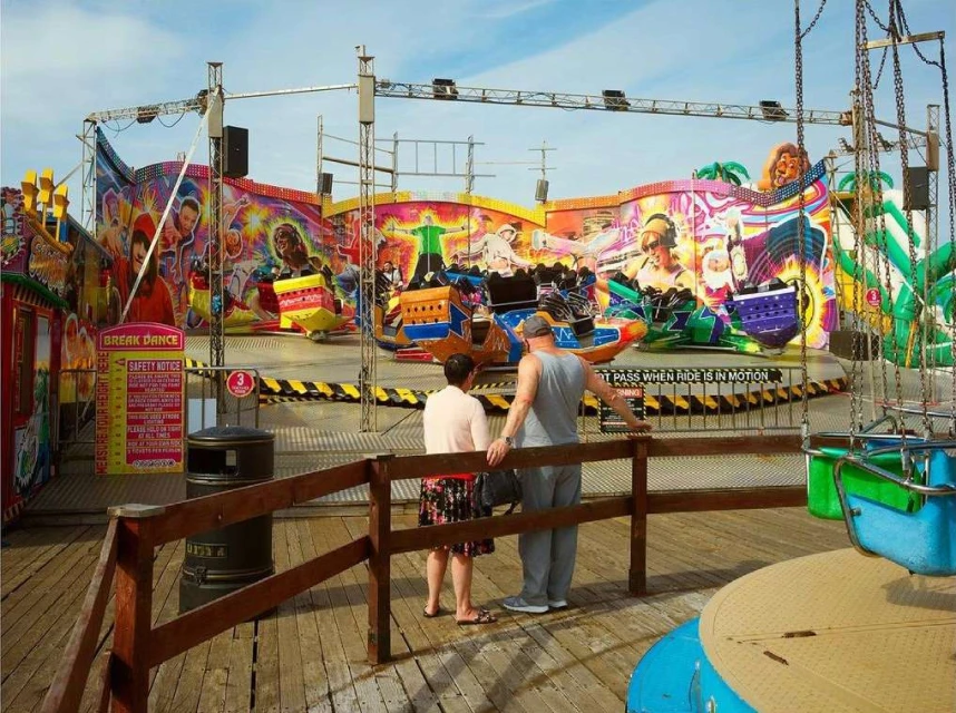 A vibrant amusement park ride spins with riders as two people observe it from a wooden boardwalk under a sunny sky.