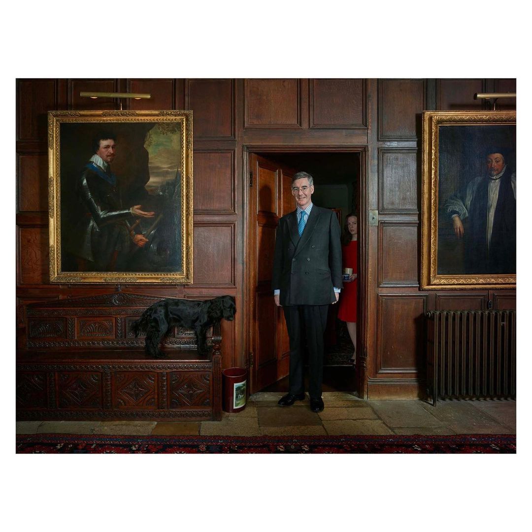 Full shot of a man in a suit standing in a wood-paneled room next to a black dog on a carved wooden chest with two large paintings.