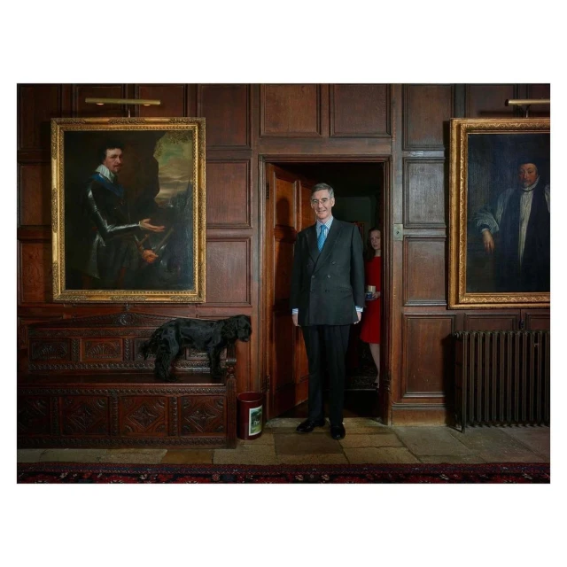 Full shot of a man in a suit standing in a wood-paneled room next to a black dog on a carved wooden chest with two large paintings.
