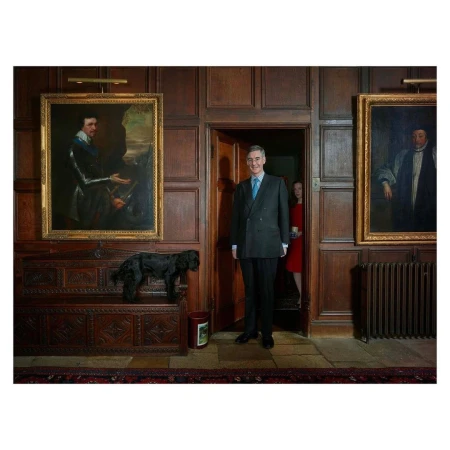 Full shot of a man in a suit standing in a wood-paneled room next to a black dog on a carved wooden chest with two large paintings.