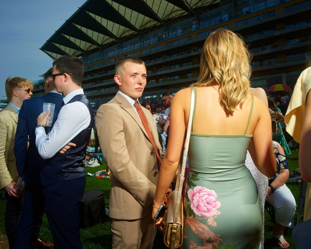 Outdoor daytime event features a man in a suit, a woman in a floral dress, and spectators at an outdoor stadium or racetrack.