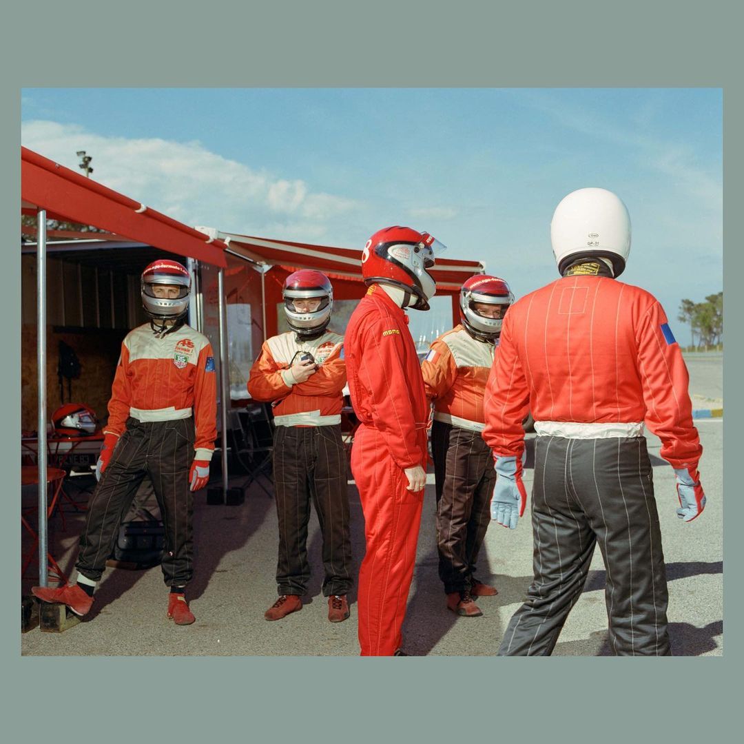 Five racecar drivers in red and white racing suits and helmets stand near a red structure under a blue sky on a racetrack.