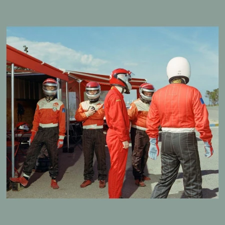 Five racecar drivers in red and white racing suits and helmets stand near a red structure under a blue sky on a racetrack.