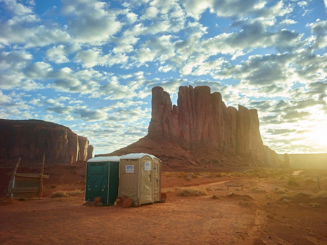 Monument Valley, Utah, vista with red rock formations, blue sky with fluffy clouds, and portable restrooms in the foreground.