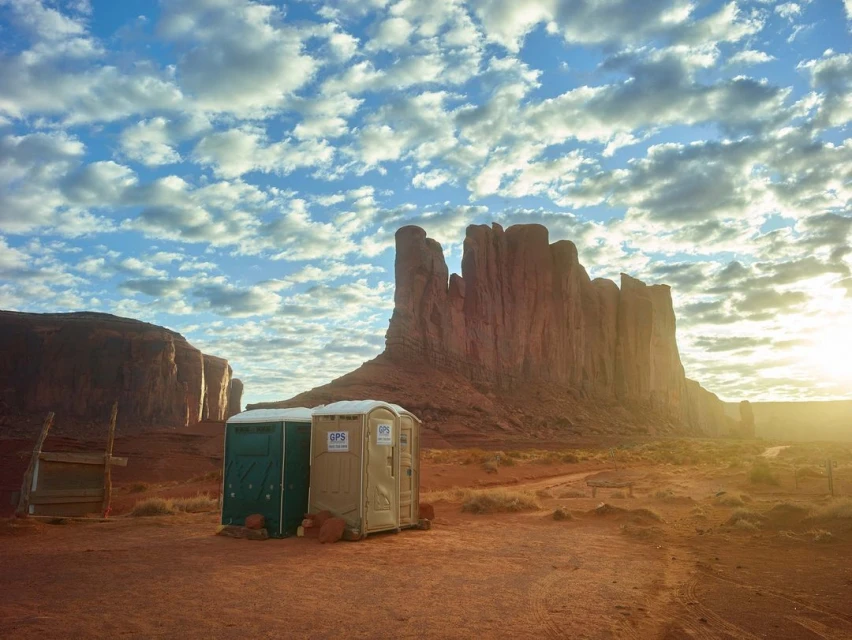 Monument Valley, Utah, vista with red rock formations, blue sky with fluffy clouds, and portable restrooms in the foreground.