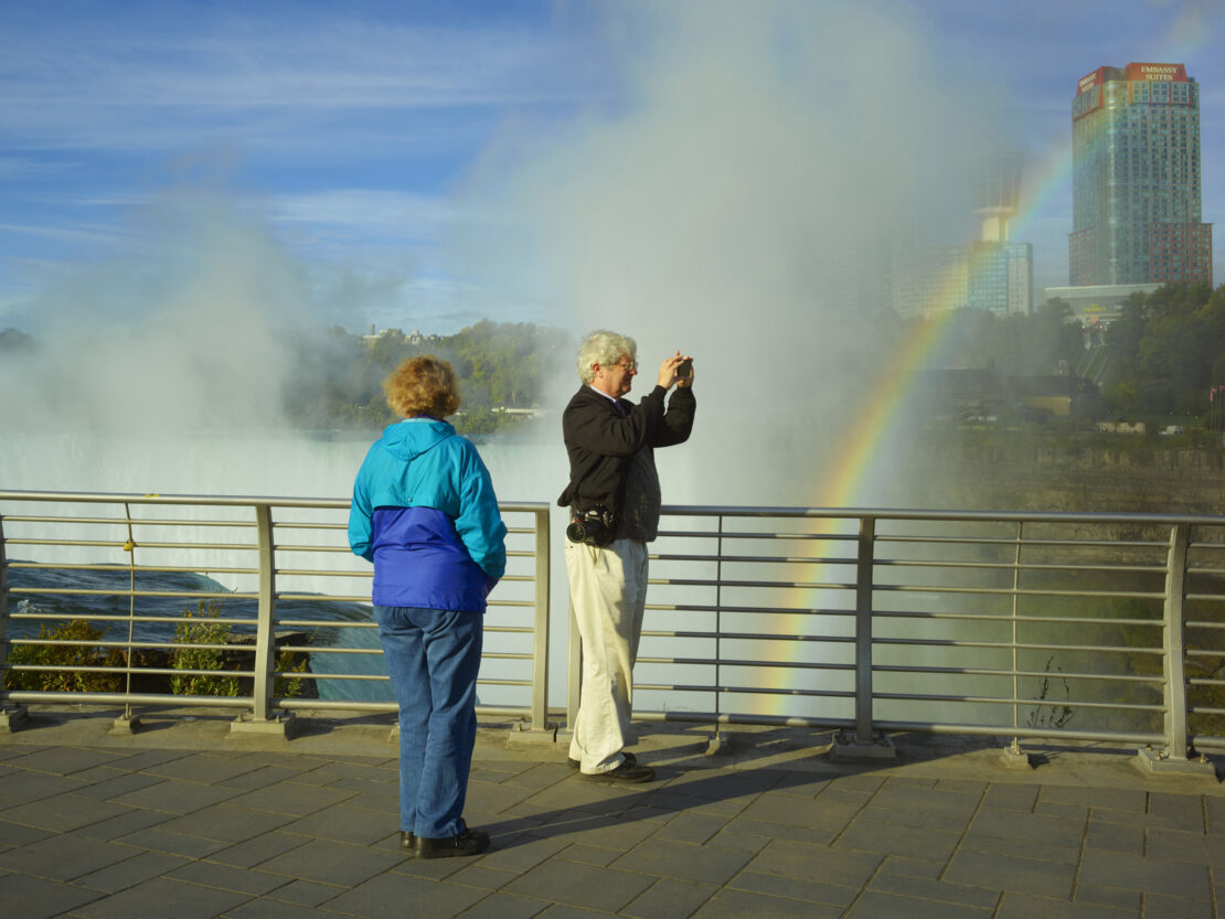Two tourists admire a rainbow at Niagara Falls, with a high-rise building and mist in the background on a sunny day, shot at eye-level.