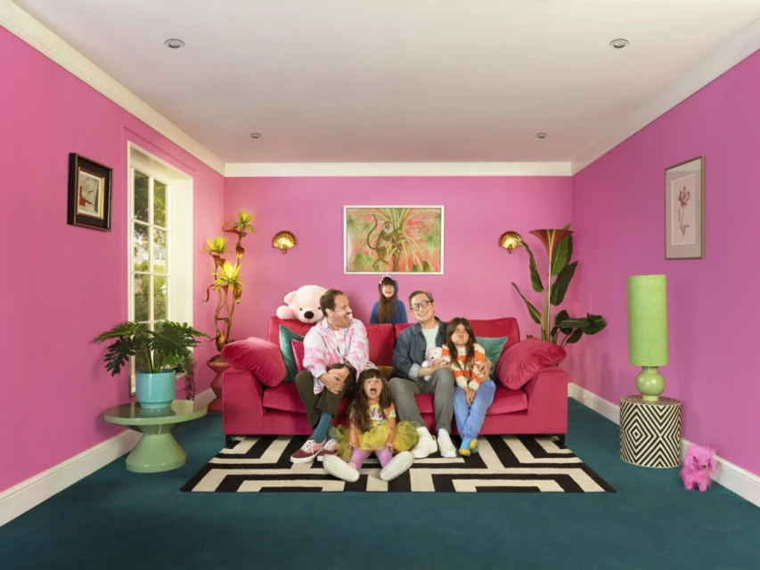 Family of five poses on a pink couch in a bright, colorful living room with teal carpet, pink walls, and natural light from a window.