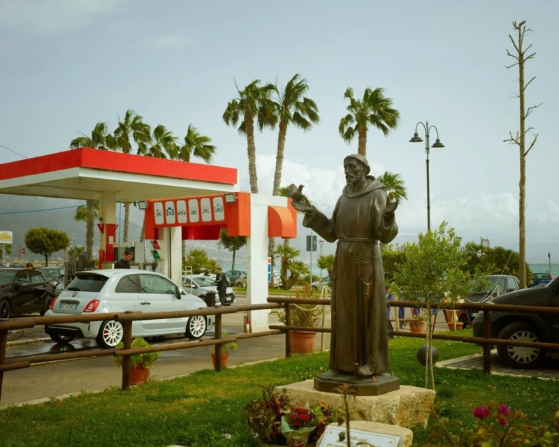 Bronze statue of Saint Francis holding a bird stands in front of a gas station and palm trees on a sunny day in an outdoor setting.