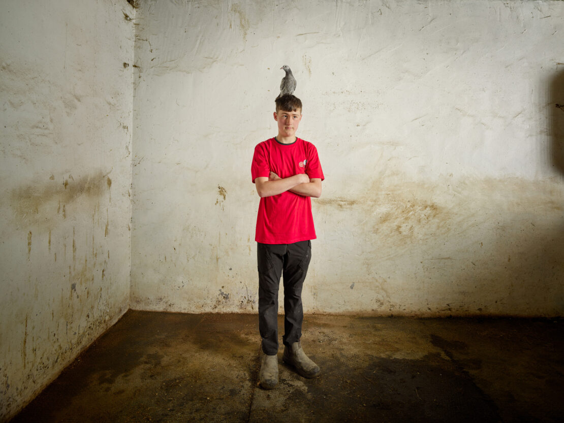 Teenager standing in concrete room with a pigeon perched atop his head, looking at the camera with arms crossed.