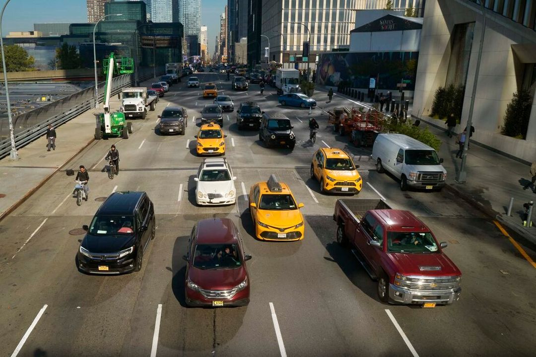 Urban street scene with multiple cars, including yellow taxis, driving on a sunny day in a city with tall buildings in the background.