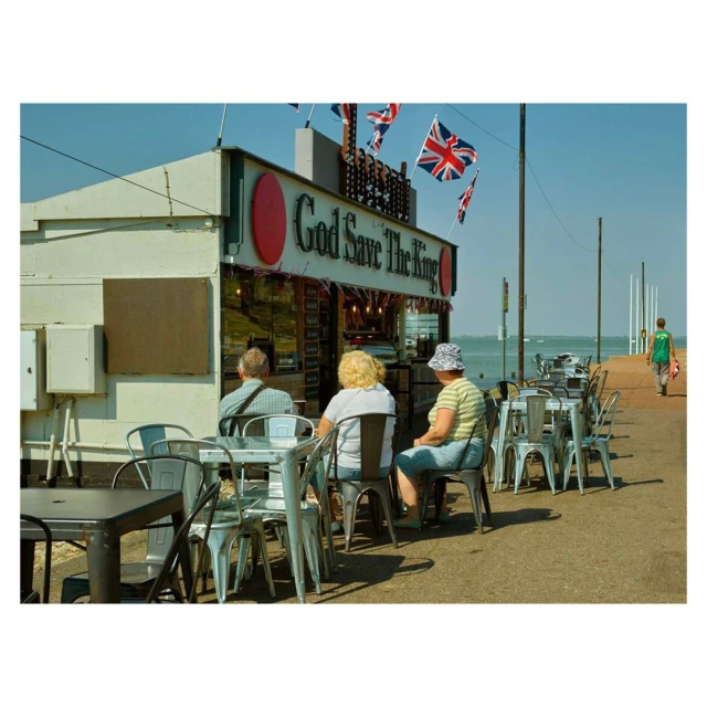 Outdoor cafe scene featuring three people seated at tables near a building adorned with British flags and 'God Save The King' signage.