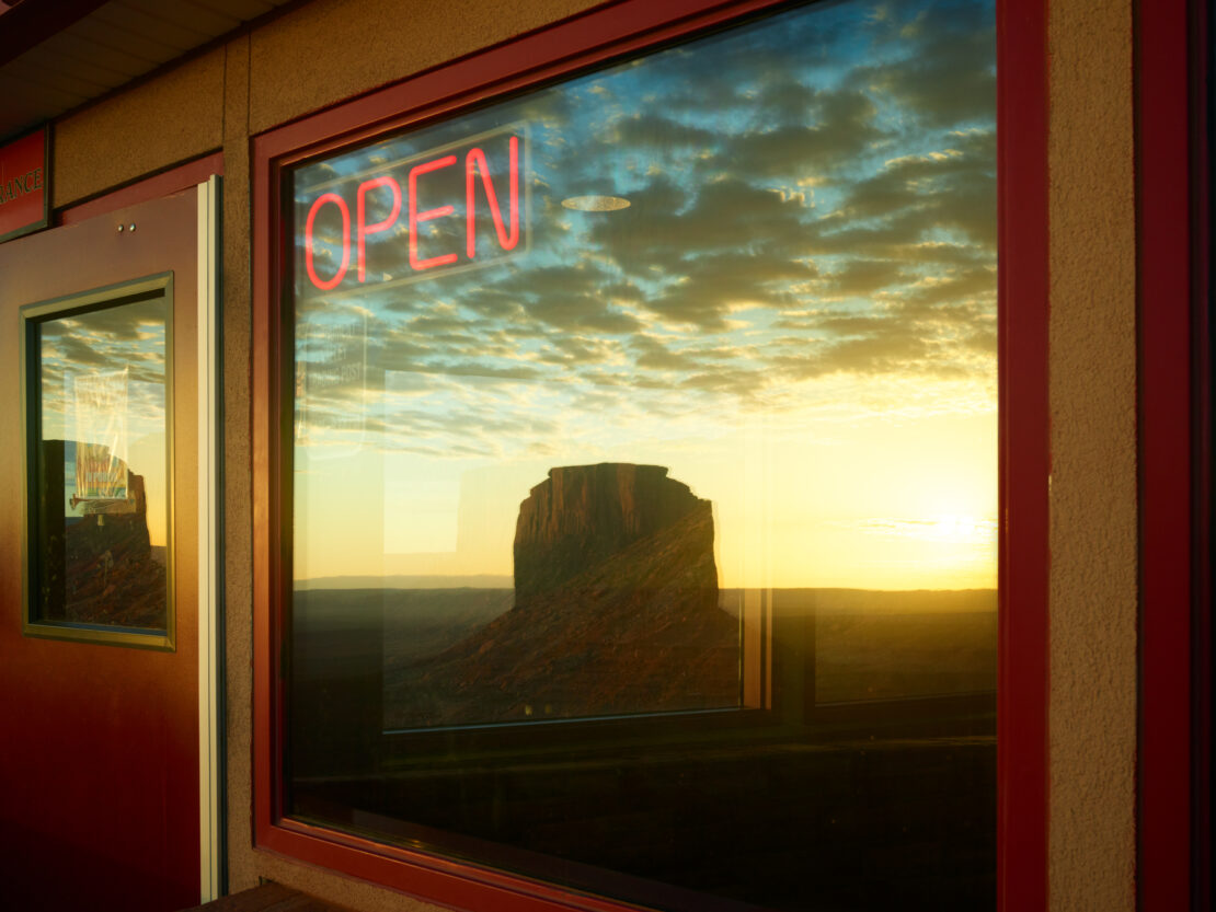 Desert butte reflected in a window with a red 'OPEN' neon sign, indicating a business is open for service in the Arizona landscape.