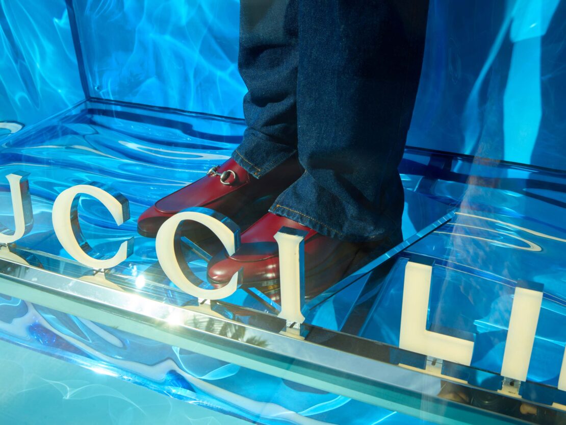 Close-up on a fashion display featuring Gucci loafers and blue jeans, set against a vibrant blue backdrop with light reflections.