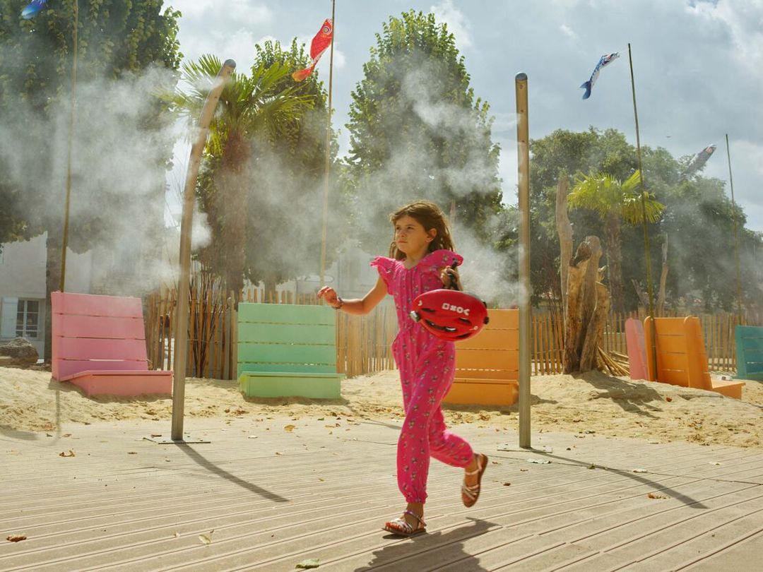 Young girl in a pink jumpsuit runs on a boardwalk with a red helmet, mist surrounds her in a park with palm trees.