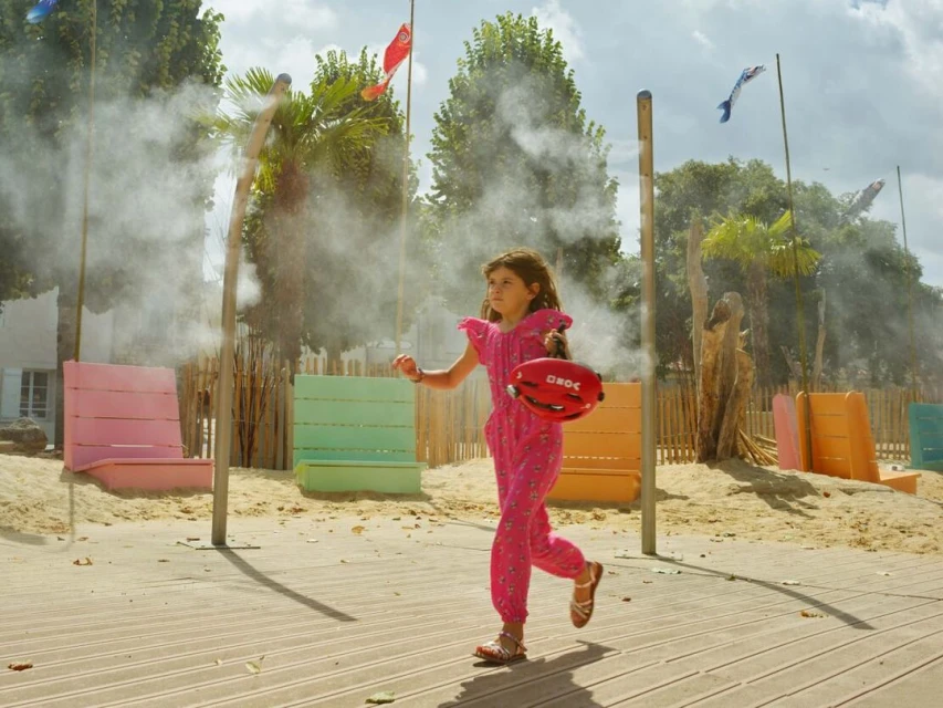 Young girl in a pink jumpsuit runs on a boardwalk with a red helmet, mist surrounds her in a park with palm trees.