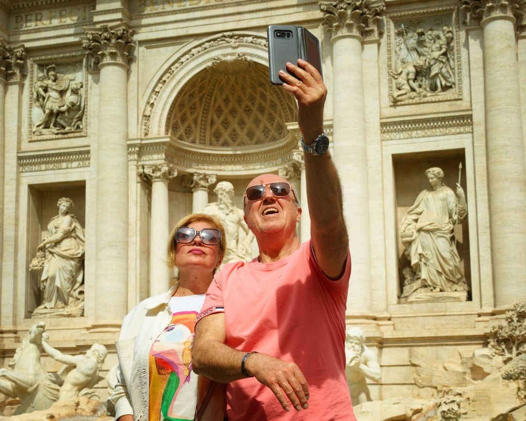 Couple takes selfie at Trevi Fountain in Rome. A man holds a phone to capture the moment. Classic architecture and white stone sculptures.